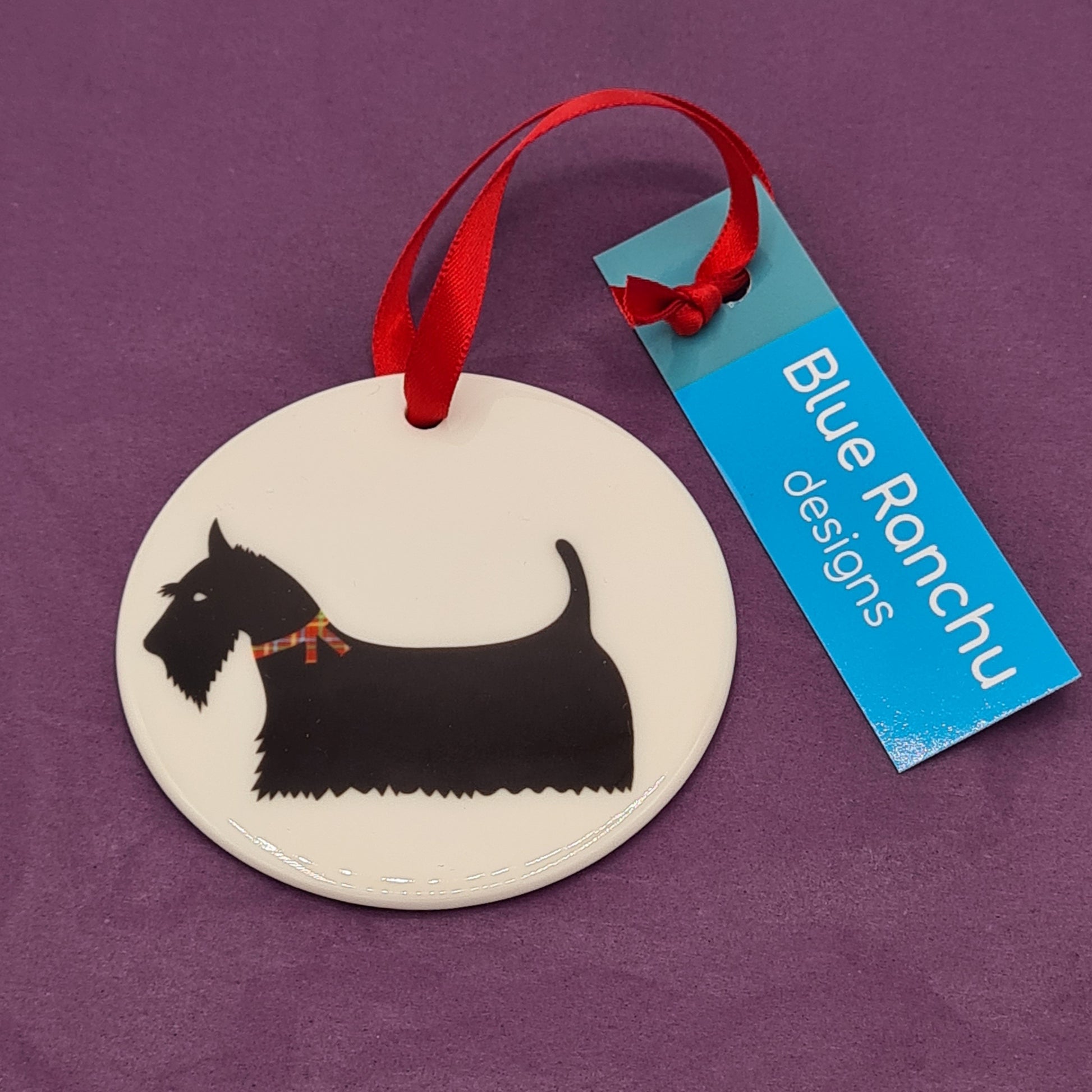 Circular white ceramic hanging decoration featuring a print of a black Scottish Terrier (Scottie dog) wearing a tartan collar around its neck. There is a red ribbon attached for hanging and a label saying "Blue Ranchu Designs"