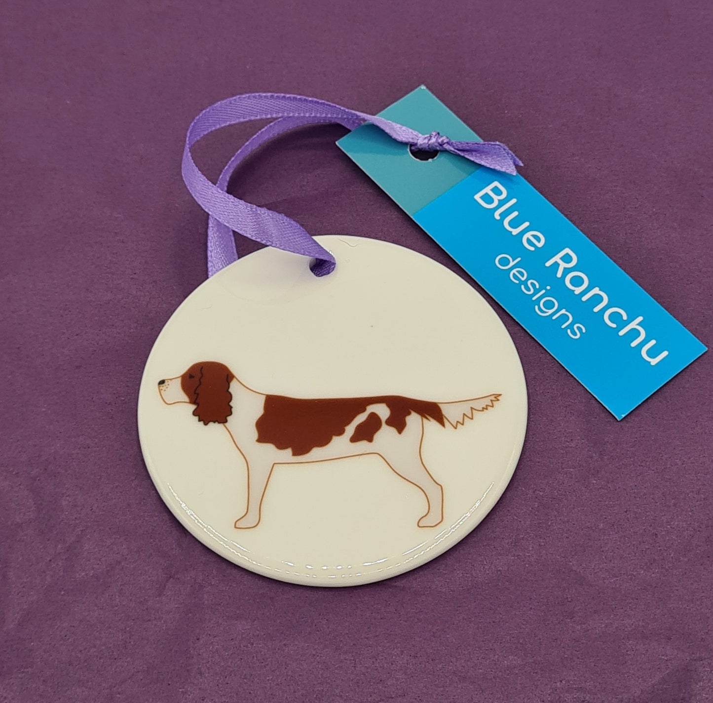 Circular white ceramic hanging decoration featuring a print of a brown and white Springer Spaniel. There is a lilac ribbon attached for hanging and a label saying "Blue Ranchu Designs".