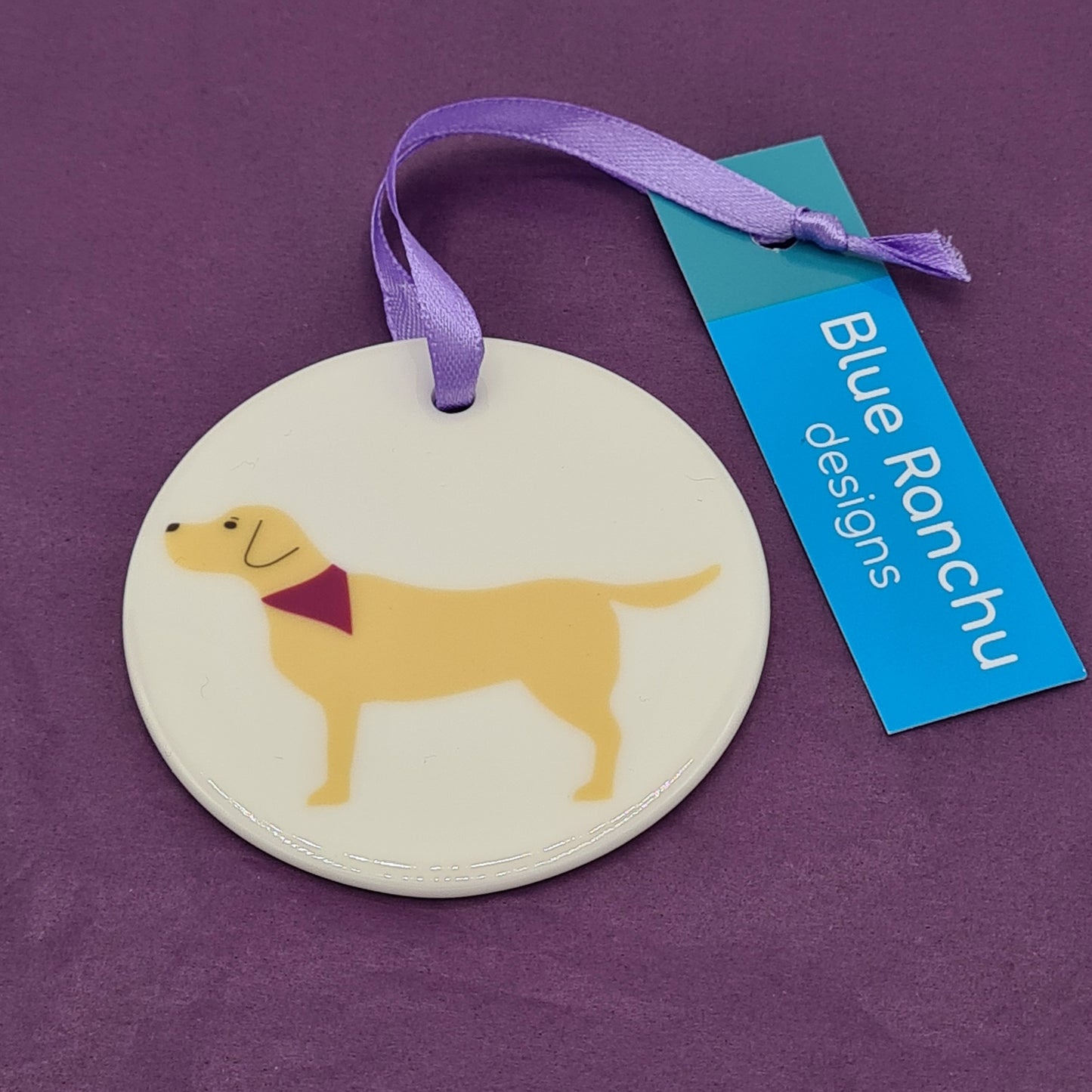 Circular white ceramic hanging decoration featuring a print of a yellow Labrador wearing a pink bandana around its neck. There is a lilac ribbon attached for hanging and a label saying "Blue Ranchu Designs".