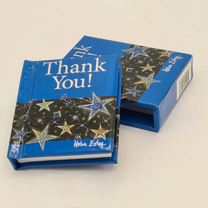 Little blue book entitled "Thank You!" with the title written in silver text. The book has its own sleeve and is shown sitting at an angle on top of the sleeve with the sleeve partially visible underneath. Both the sleeve and the book have illustrations of different coloured stars half way down the page.