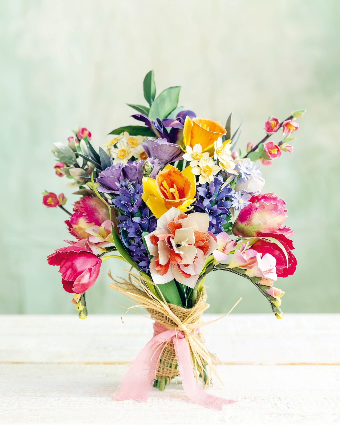 A vibrant spring flower bouquet featuring tulips, daisies, hyacinths, and roses in pink, purple, yellow, and white, wrapped in a natural jute twine with a pink ribbon, set against a soft green background. 