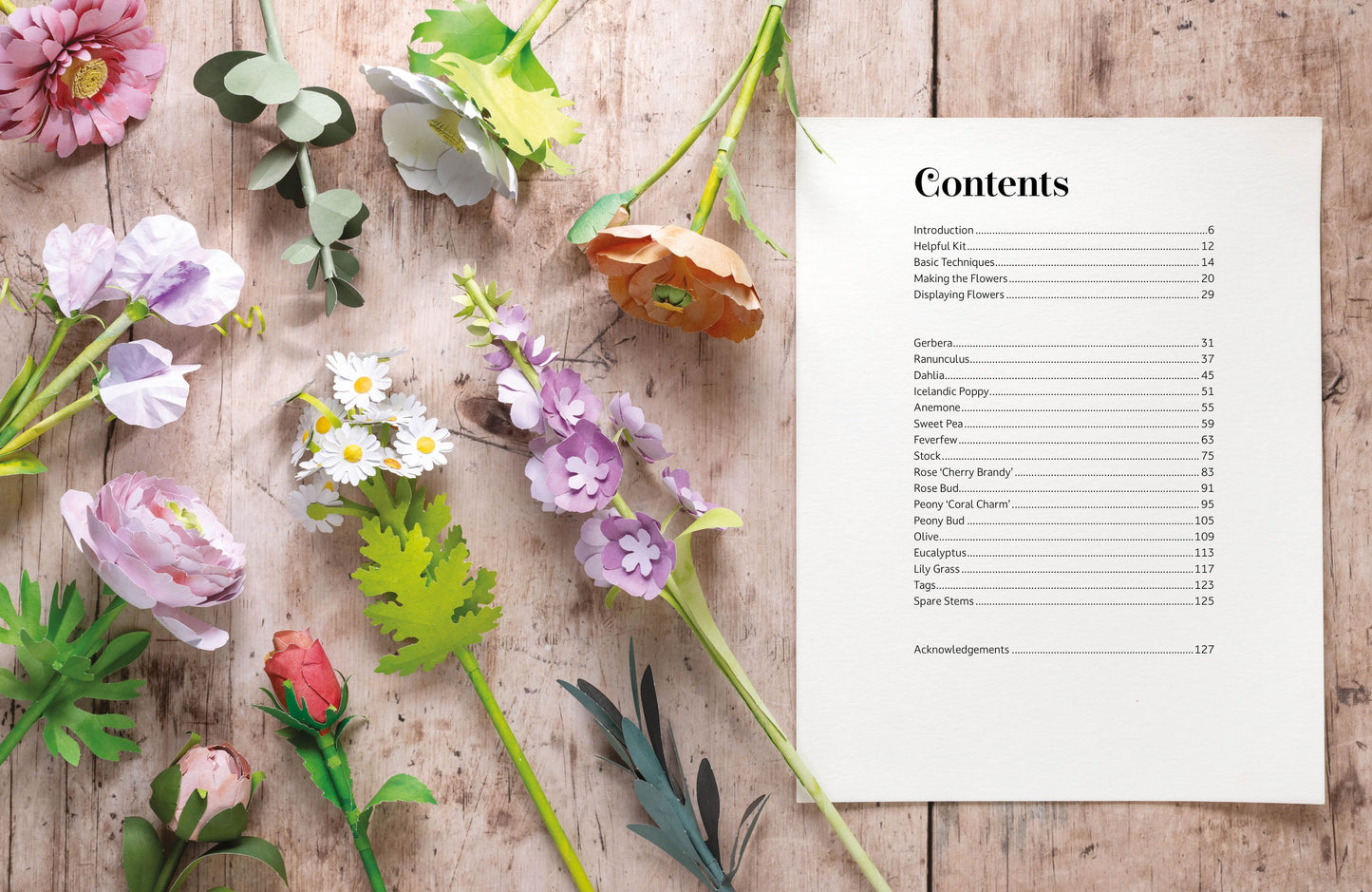 A printed table of contents page with black text on a white sheet, arranged on a rustic wooden surface alongside colorful artificial paper flowers and green leaves.