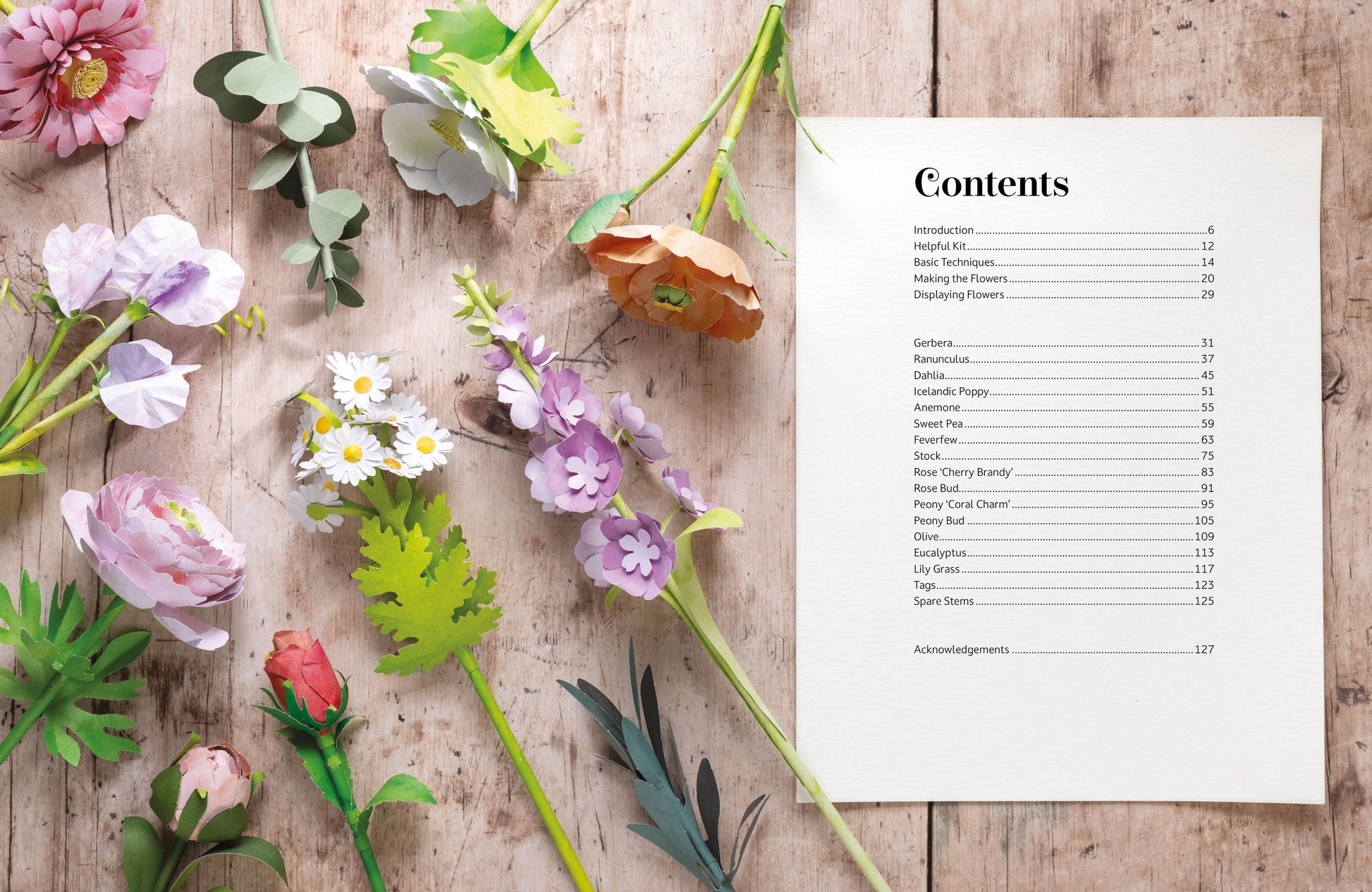 A printed table of contents page with black text on a white sheet, arranged on a rustic wooden surface alongside colorful artificial paper flowers and green leaves.