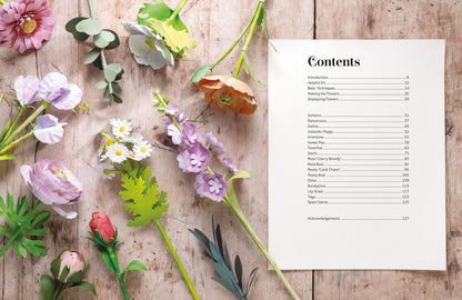 A printed table of contents page with black text on a white sheet, arranged on a rustic wooden surface alongside colorful artificial paper flowers and green leaves.