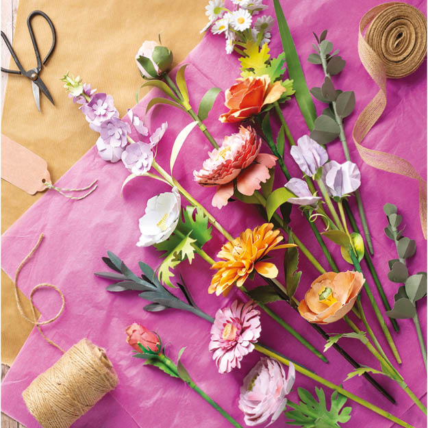 A styled flat-lay of artificial flower stems arranged on bright pink paper, with scissors, twine, ribbon, and kraft paper visible around them. The flowers include orange, pink, purple, white, and green tones.
