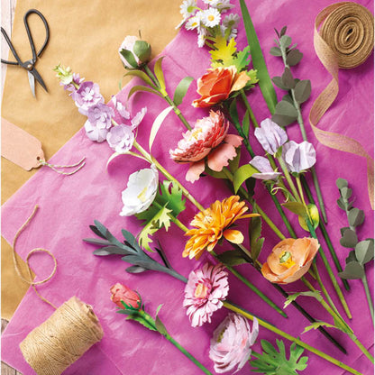A styled flat-lay of artificial flower stems arranged on bright pink paper, with scissors, twine, ribbon, and kraft paper visible around them. The flowers include orange, pink, purple, white, and green tones.