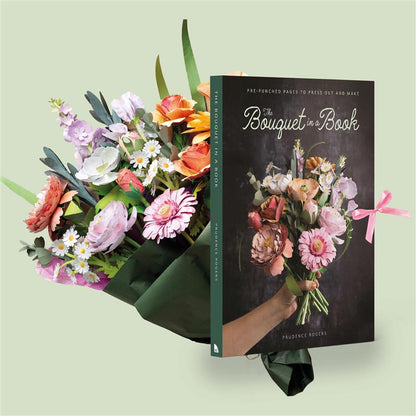A book titled "The Bouquet in a Book" by Prudence Rogers, shown standing upright next to a colourful floral bouquet wrapped in dark green paper. The book cover features a bouquet illustration and a hand holding flowers, set against a pale sage green background.