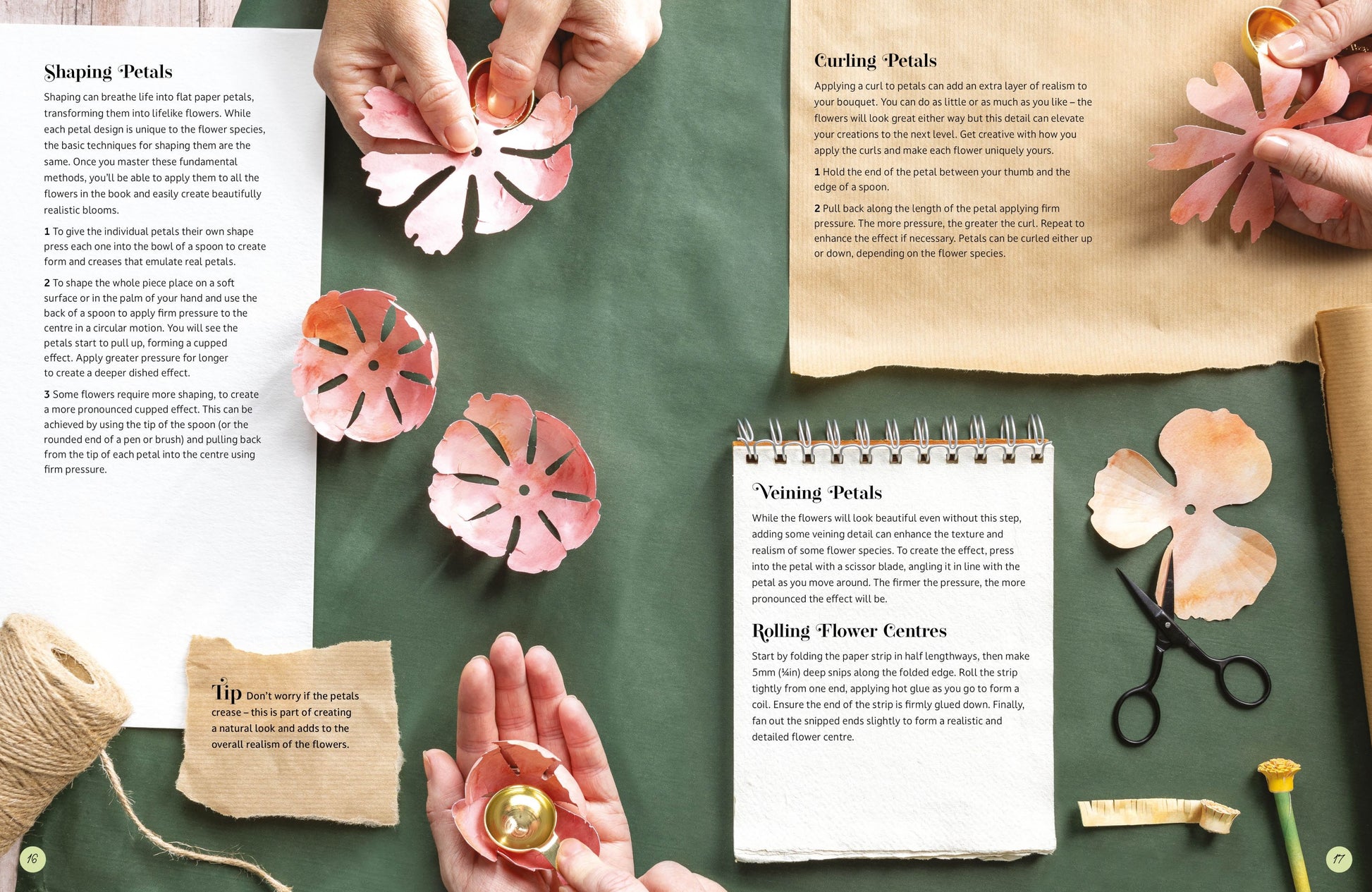 A styled flat-lay of a paper flower craft kit showing hands shaping pink paper flowers, with printed instruction sheets, scissors, and other crafting tools arranged on a dark green surface.