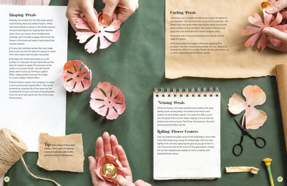 A styled flat-lay of a paper flower craft kit showing hands shaping pink paper flowers, with printed instruction sheets, scissors, and other crafting tools arranged on a dark green surface.