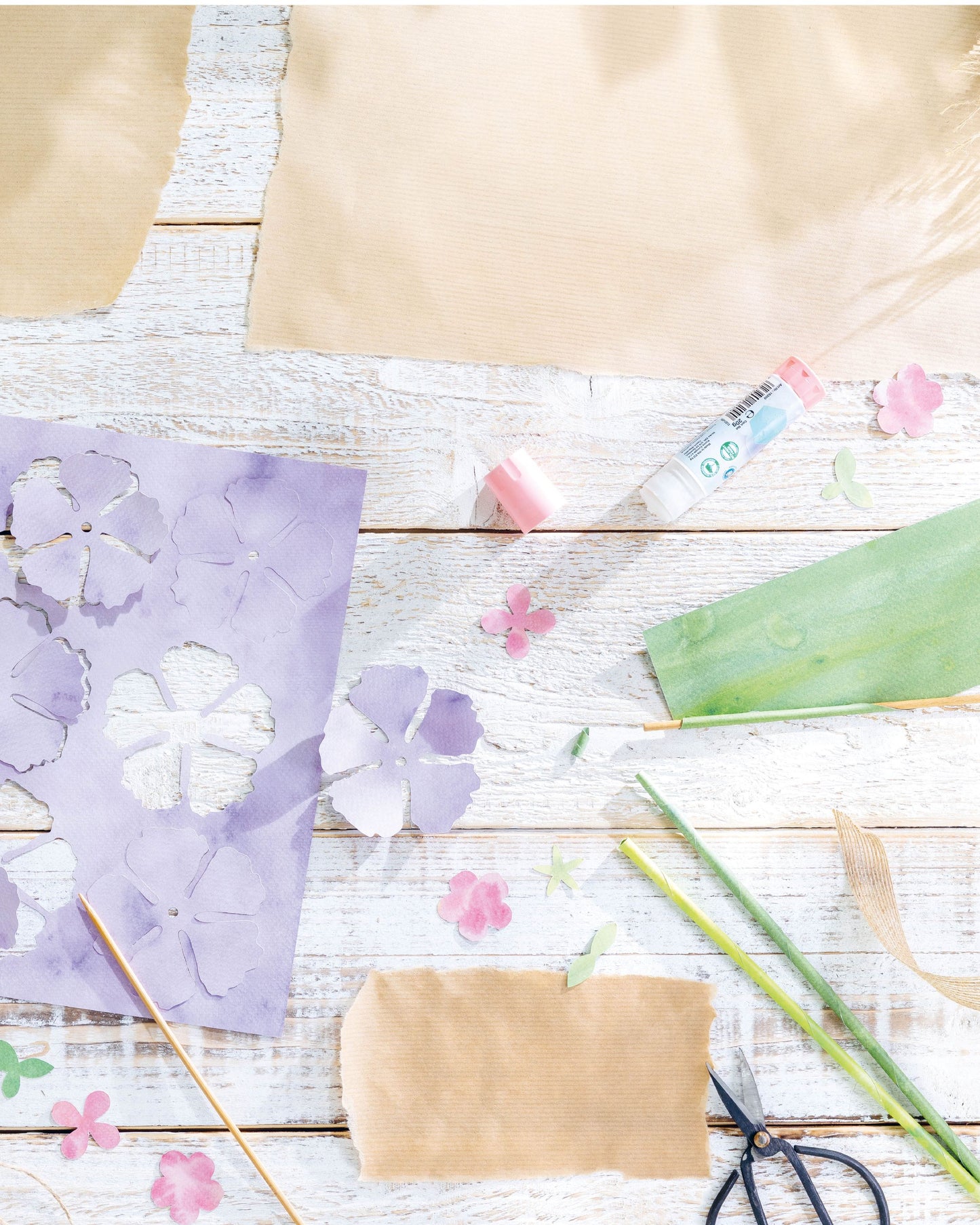 A styled flat-lay of craft supplies on a white wood surface, including purple flower cutouts on paper, brown paper sheets, a green leaf-shaped piece, pink flower petals, scissors, green stems, and a small tube of glue.