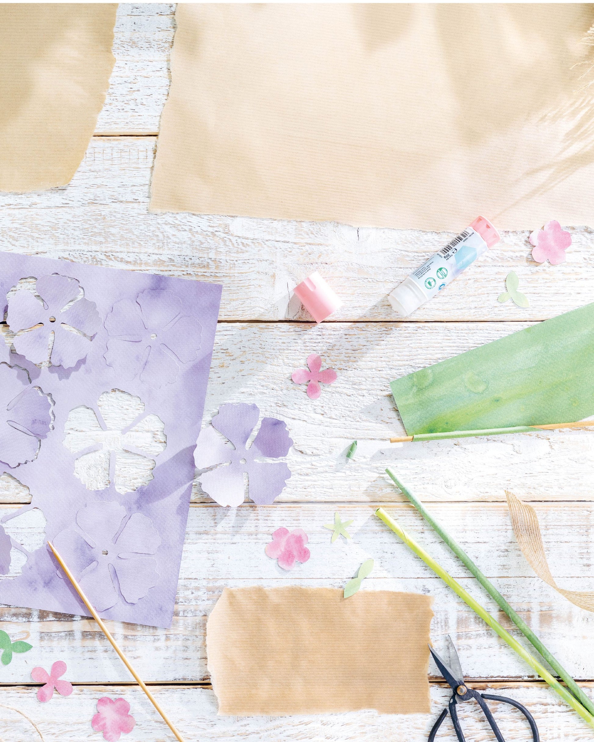 A styled flat-lay of craft supplies on a white wood surface, including purple flower cutouts on paper, brown paper sheets, a green leaf-shaped piece, pink flower petals, scissors, green stems, and a small tube of glue.