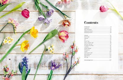 A printed contents page with a list of sections and page numbers, placed on a white wood surface and surrounded by scattered tulips, hyacinths, and other flowers.