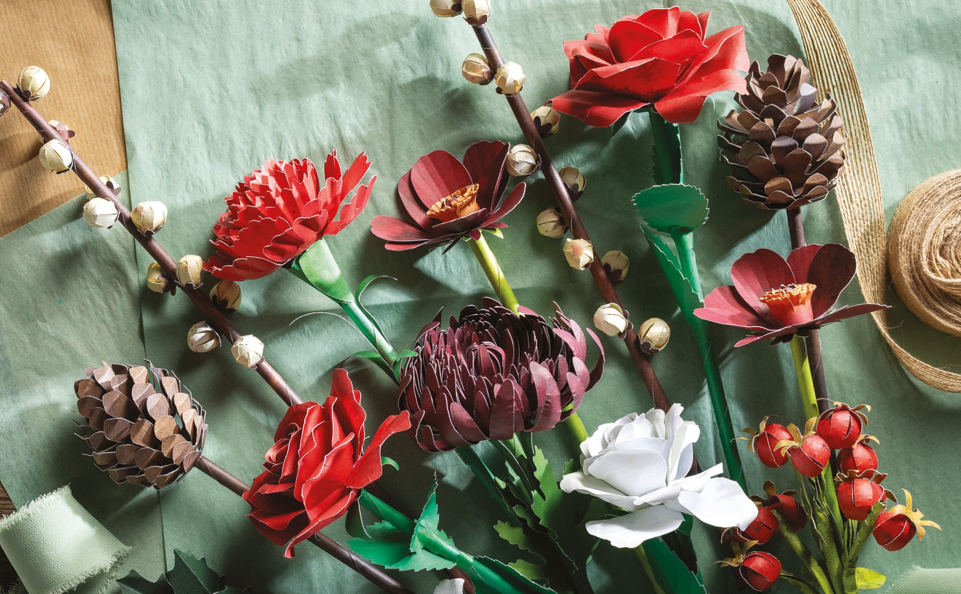 A styled flat-lay of paper flowers and botanical elements arranged on a textured sage green fabric background, including red and burgundy blossoms, a white rose, pinecones, berries, and small beige buds.