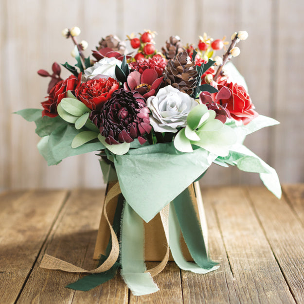 A decorative artificial paper floral arrangement in a square container, featuring red and burgundy faux roses, a white rose, green leaves, pinecones, and ribbon-wrapped stems, set on a wooden tabletop with a soft neutral background.