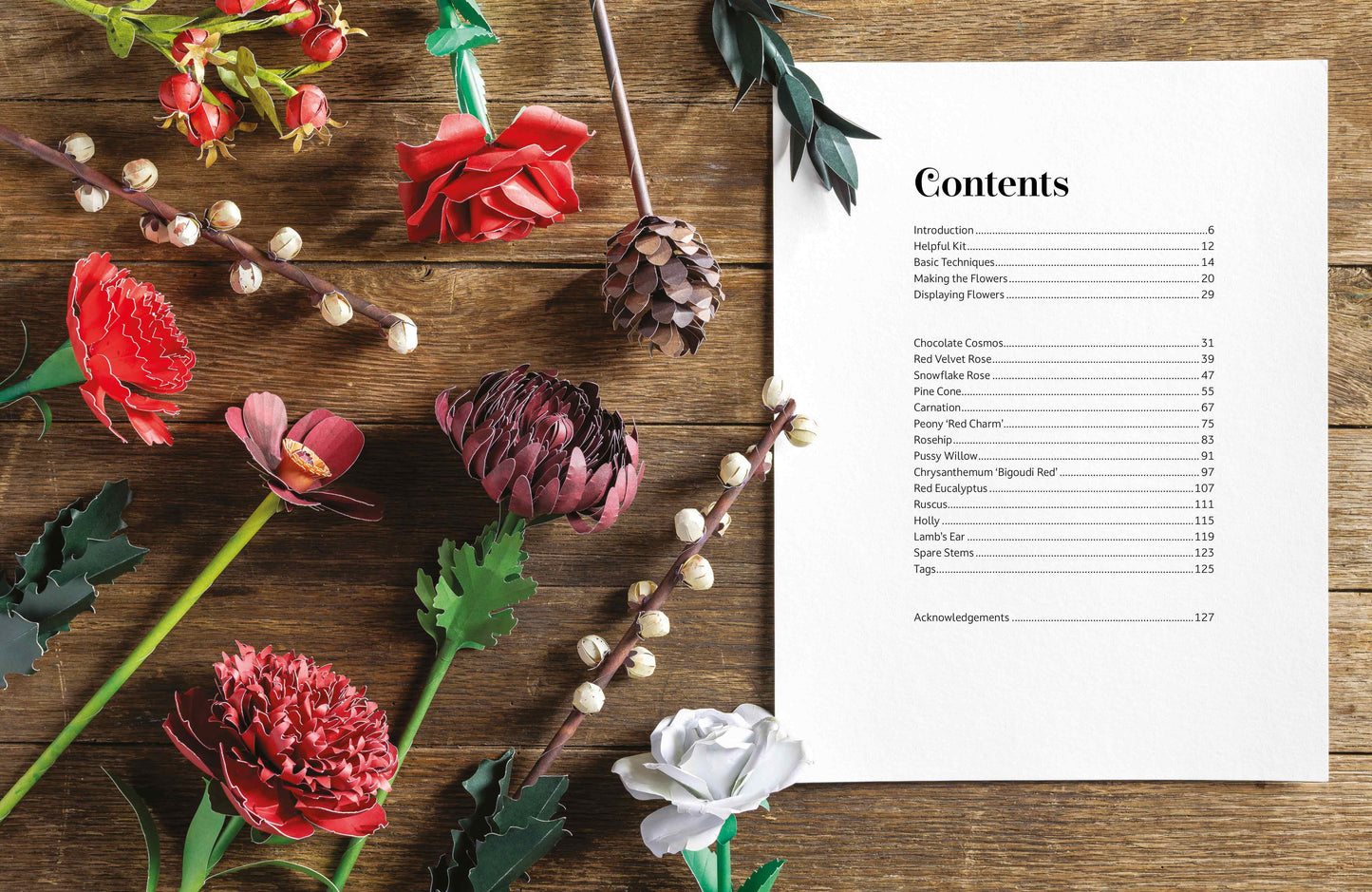 A printed contents page with black text on a white sheet, arranged on a rustic wooden surface alongside decorative red and white flowers, green leaves, and small white berries.