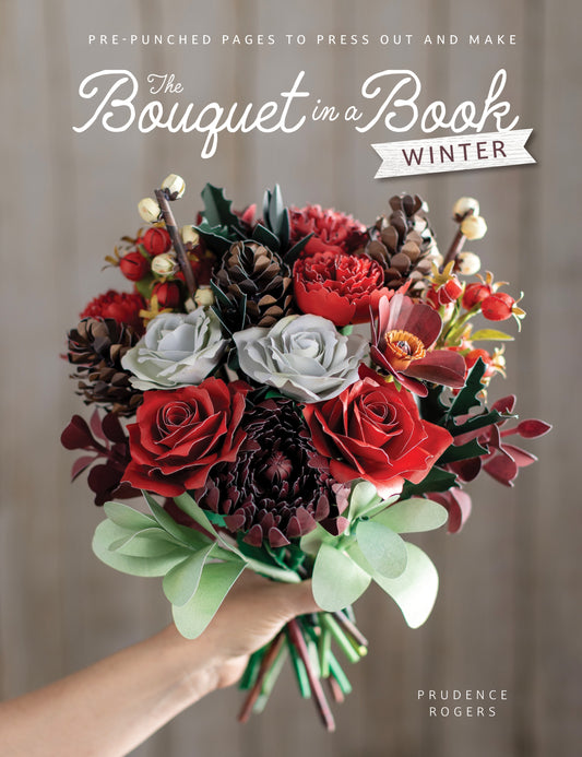A book cover showing a hand holding a large, intricate paper bouquet of winter flowers and foliage, featuring red roses, white and gray roses, pinecones, and green leaves. The background is a soft, neutral grey-brown wood texture, with the title 'The Bouquet in a Book: Winter' and the author name 'Prudence Rogers' printed on the cover.
