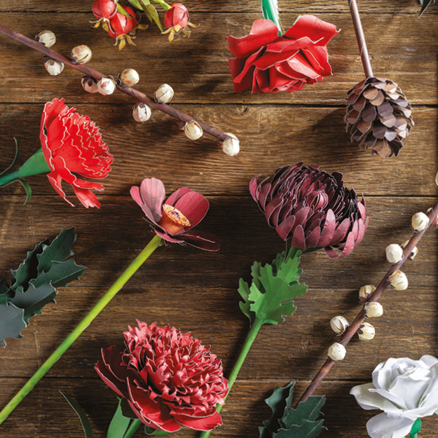 A styled flat-lay of artificial flowers and botanical elements arranged on a rustic dark wood surface, including red and pink blossoms, green leaves, brown branches with small white berries, a pinecone, and a white flower.