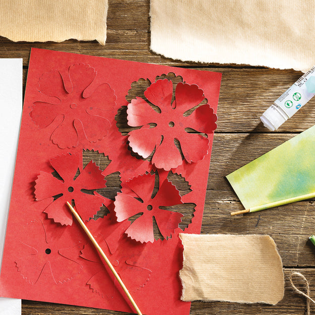 A red paper craft sheet with layered cutout floral shapes, arranged on a rustic wooden surface with paper, a wooden skewer, and a glue tube nearby.