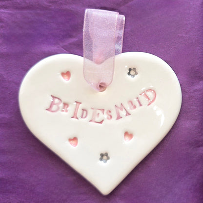 White ceramic heart with the words "Bridesmaid" and some small heart and flower shaped illustrations dotted around. There is a pale pink ribbon at the top of the heart for hanging.