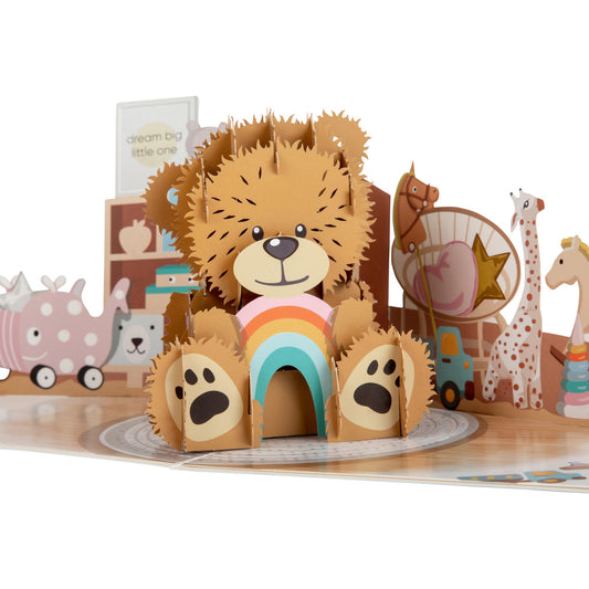 Close up of New Baby Bear pop up card showing cute brown teddy bear holding a rainbow. The bear appears to be sitting in a nursery and is surrounded by various toys.