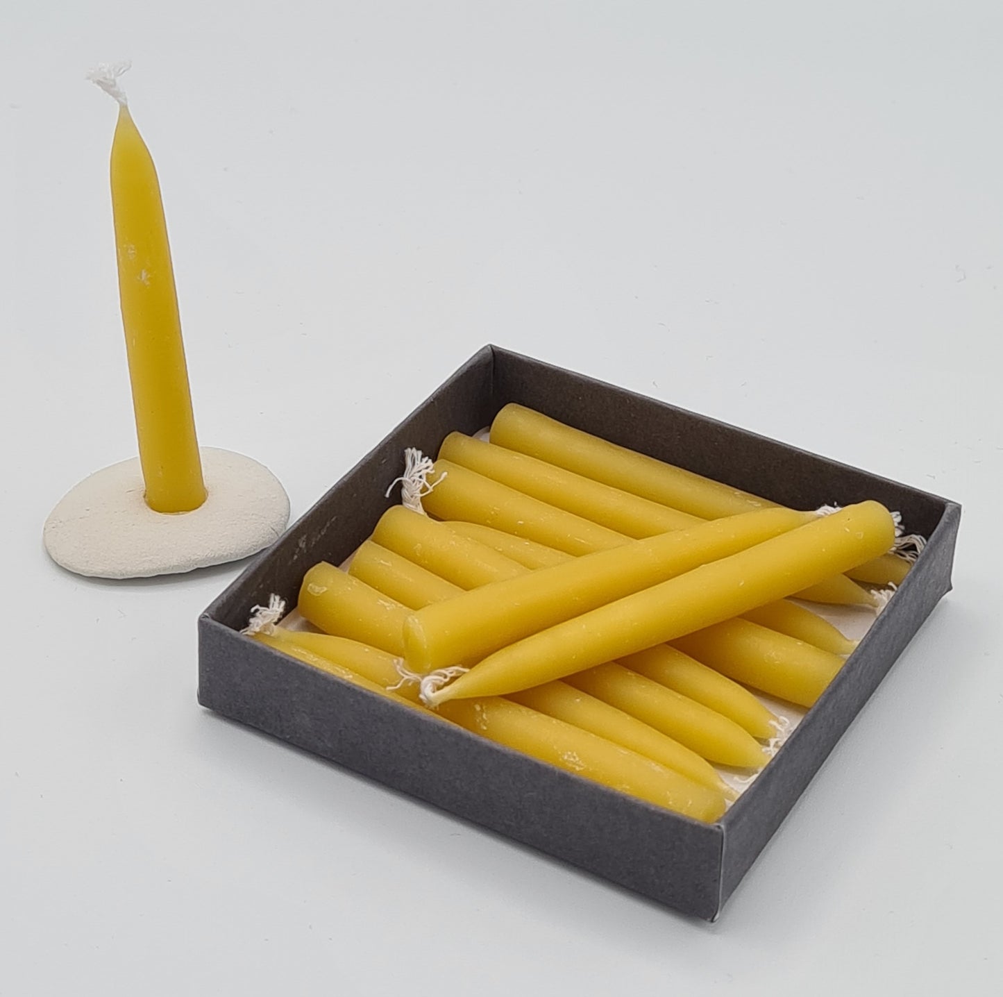 The inside of the small square box showing 11 mini yellow beeswax candles. The 12th candle is standing in the mini ceramic holder next to the box. 