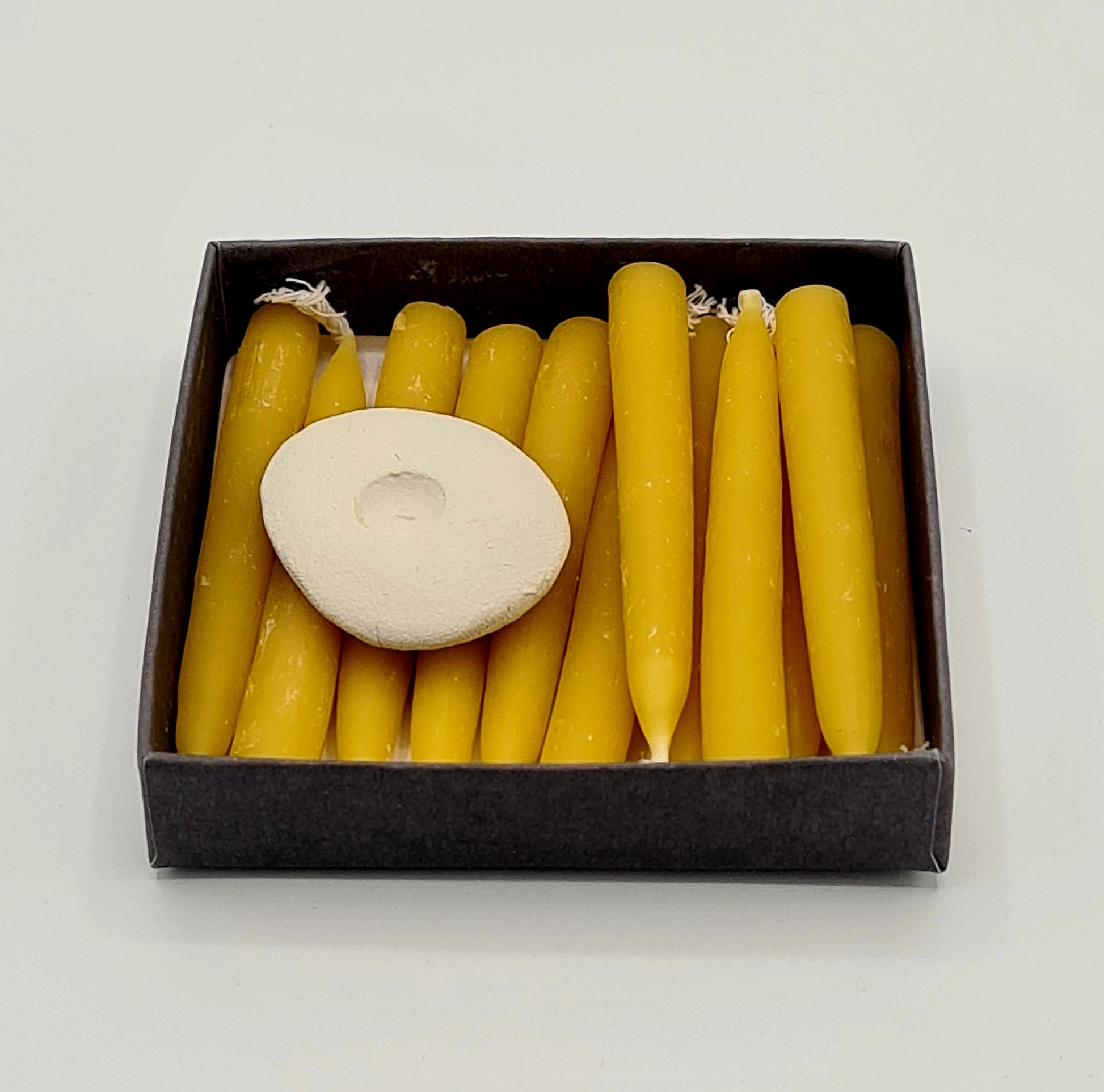 The inside of the small square box with twelve mini yellow beeswax candles and a mini ceramic candle holder.