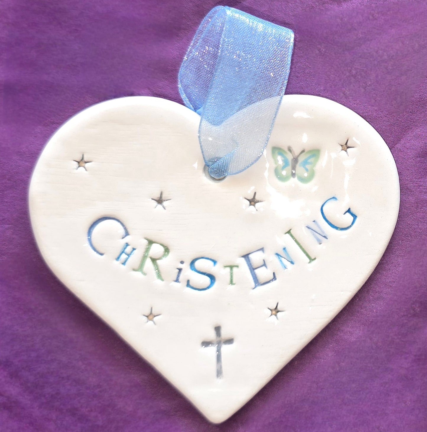 White ceramic heart with the words "Christening" in blue and some small star shapes, a blue butterfly and grey cross illustrations dotted around. There is a pale blue ribbon at the top of the heart for hanging.