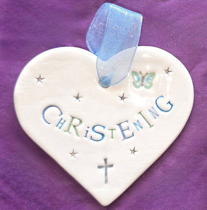 White ceramic heart with the words "Christening" in blue and some small star shapes, a blue butterfly and grey cross illustrations dotted around. There is a pale blue ribbon at the top of the heart for hanging.