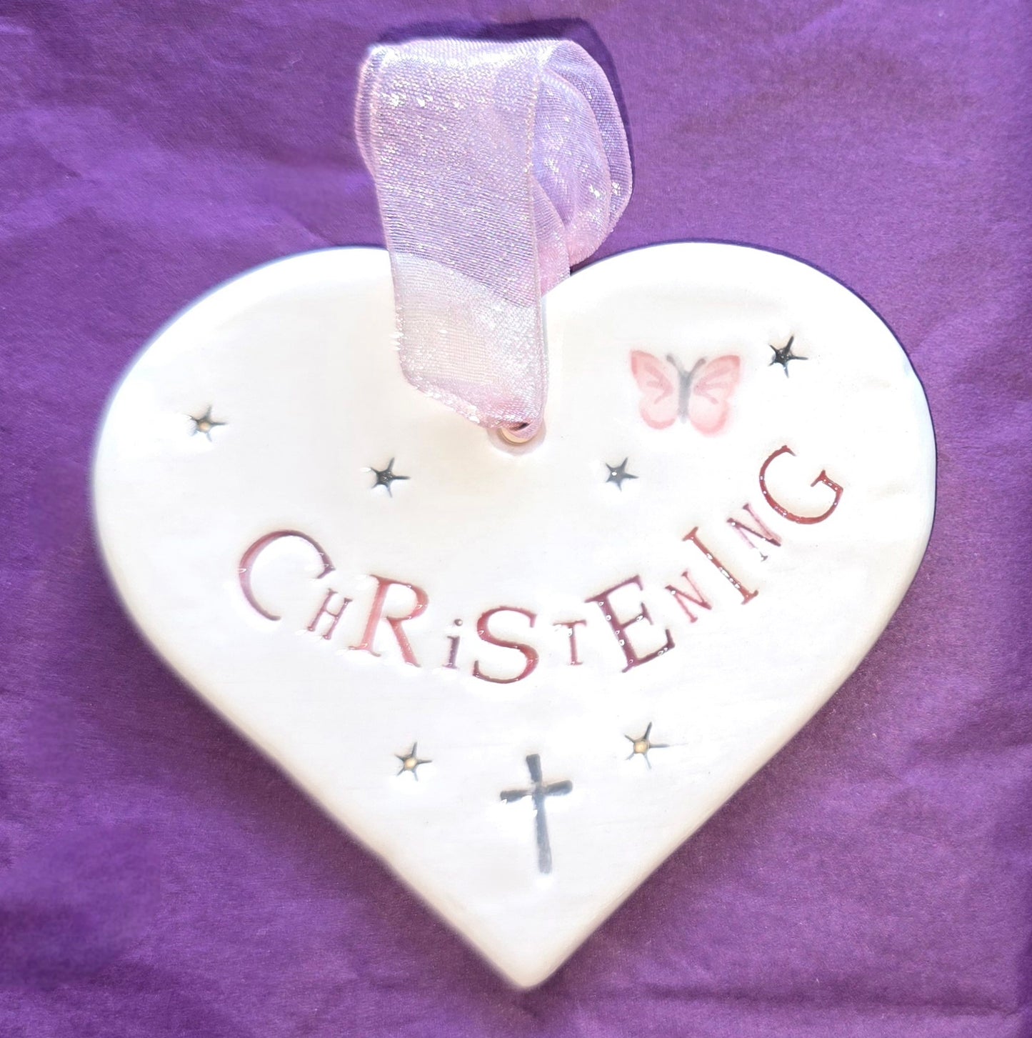 White ceramic heart with the words "Christening" in pink and some small star shapes, a pink butterfly and grey cross illustrations dotted around. There is a pale pink ribbon at the top of the heart for hanging.