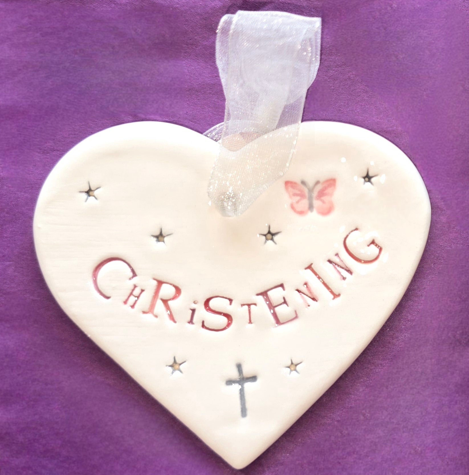 White ceramic heart with the words "Christening" in pink and some small star shapes, a pink butterfly and grey cross illustrations dotted around. There is a white ribbon at the top of the heart for hanging.