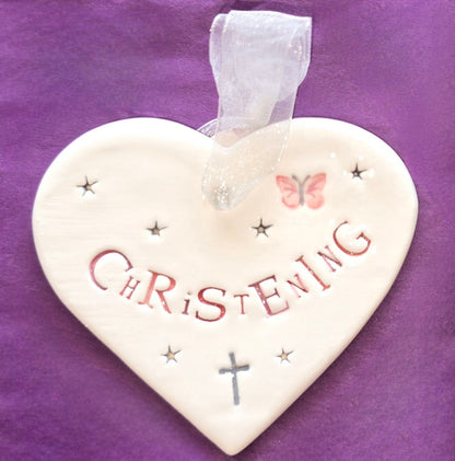 White ceramic heart with the words "Christening" in pink and some small star shapes, a pink butterfly and grey cross illustrations dotted around. There is a white ribbon at the top of the heart for hanging.