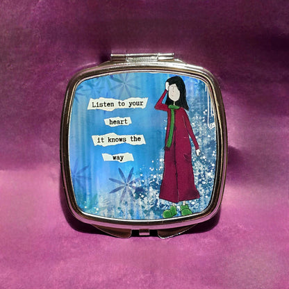 Square silver compact mirror. On the lid there is a sketch of a lady with dark hair wearing a burgundy coat with green scarf and shoes. To the left of the lady is the text "Listen to your heart it knows the way".