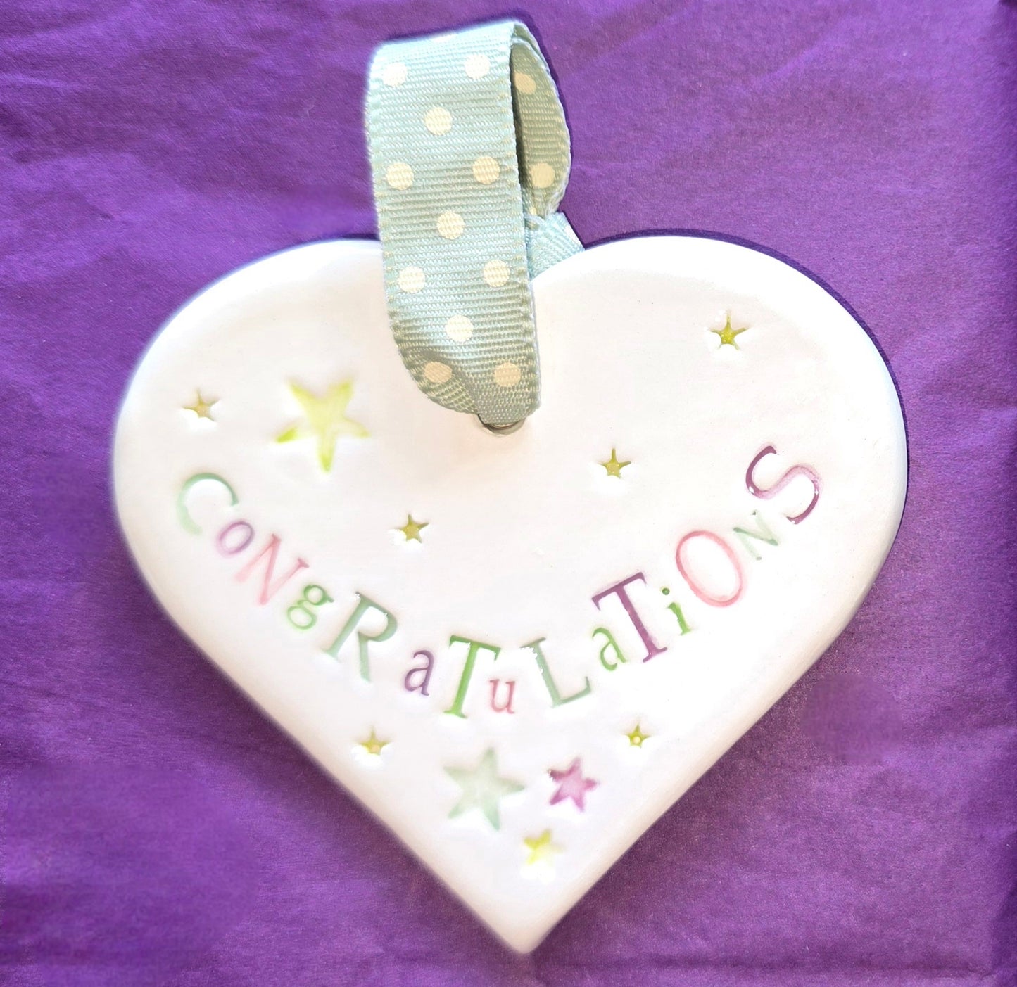 White ceramic heart with the words "Congratulations" and some small star shaped illustrations dotted around. There is a light green polka dot ribbon at the top of the heart for hanging.