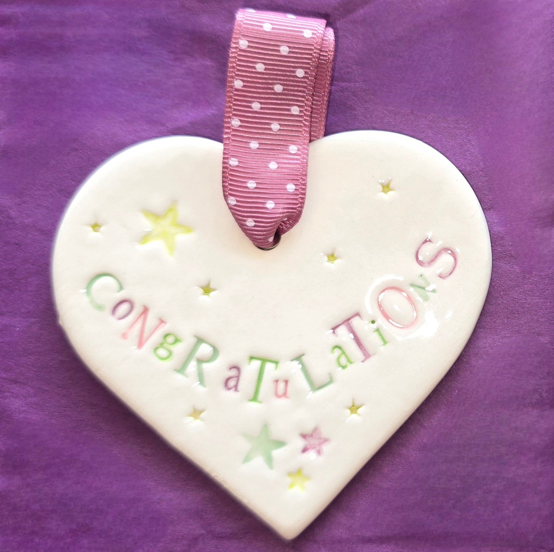 White ceramic heart with the words "Congratulations" and some small star shaped illustrations dotted around. There is a pink polka dot ribbon at the top of the heart for hanging.