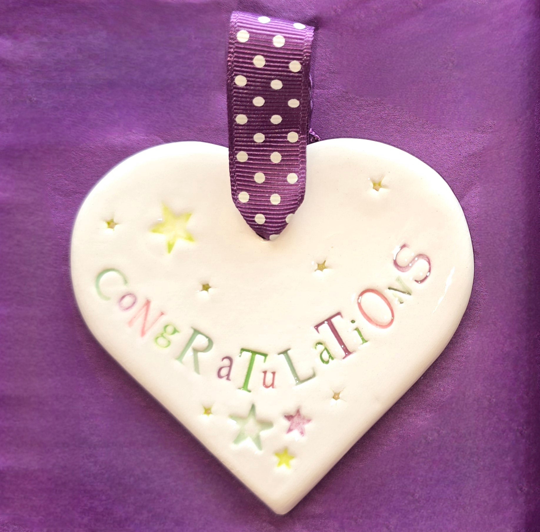 White ceramic heart with the words "Congratulations" and some small star shaped illustrations dotted around. There is a purple polka dot ribbon at the top of the heart for hanging.