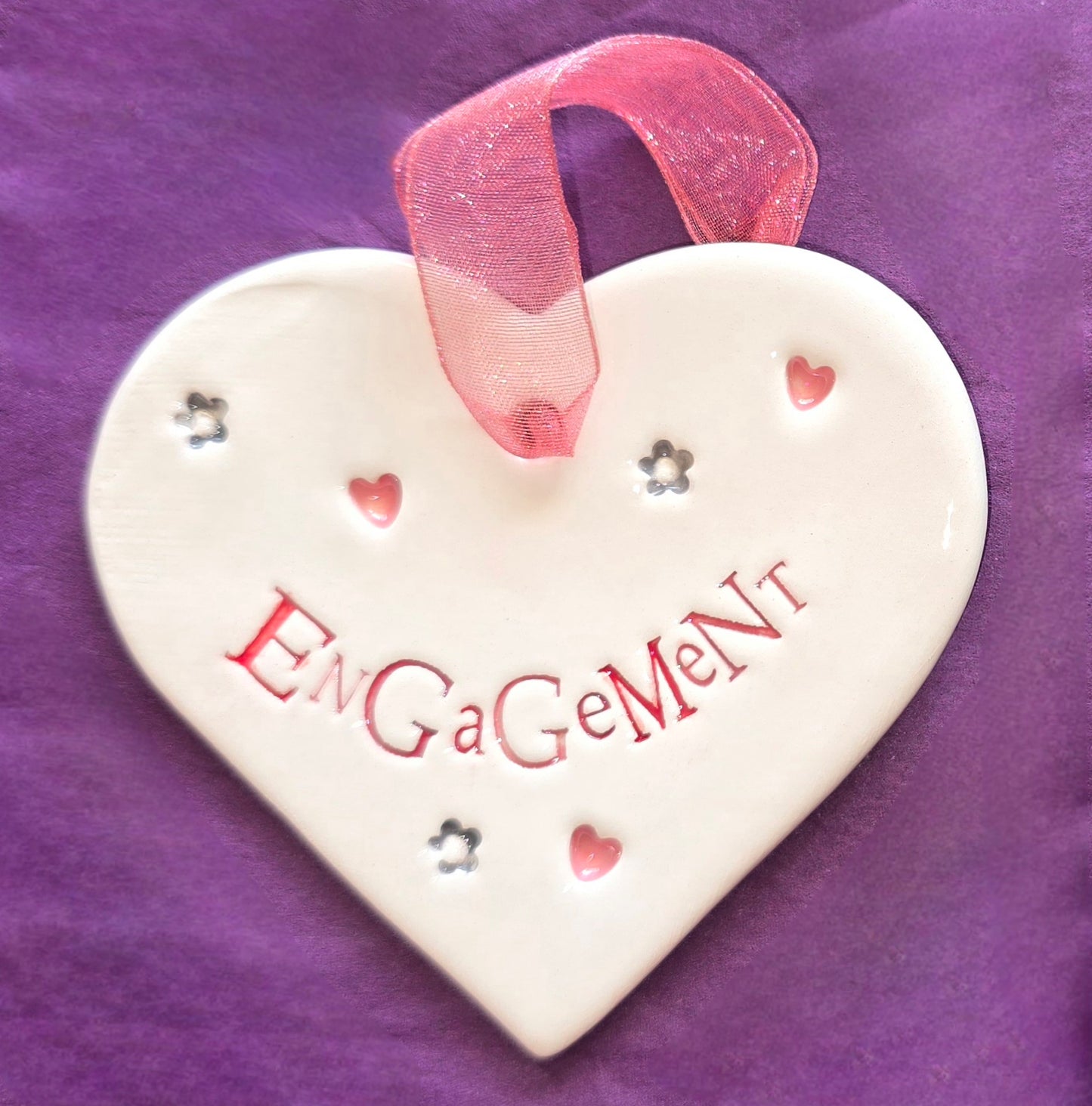 White ceramic heart with the words "Engagement" and some small heart and flower shaped illustrations dotted around. There is a pink ribbon at the top of the heart for hanging.