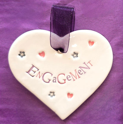 White ceramic heart with the words "Engagement" and some small heart and flower shaped illustrations dotted around. There is a purple ribbon at the top of the heart for hanging.