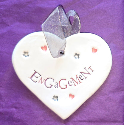 White ceramic heart with the words "Engagement" and some small heart and flower shaped illustrations dotted around. There is a silver ribbon at the top of the heart for hanging.