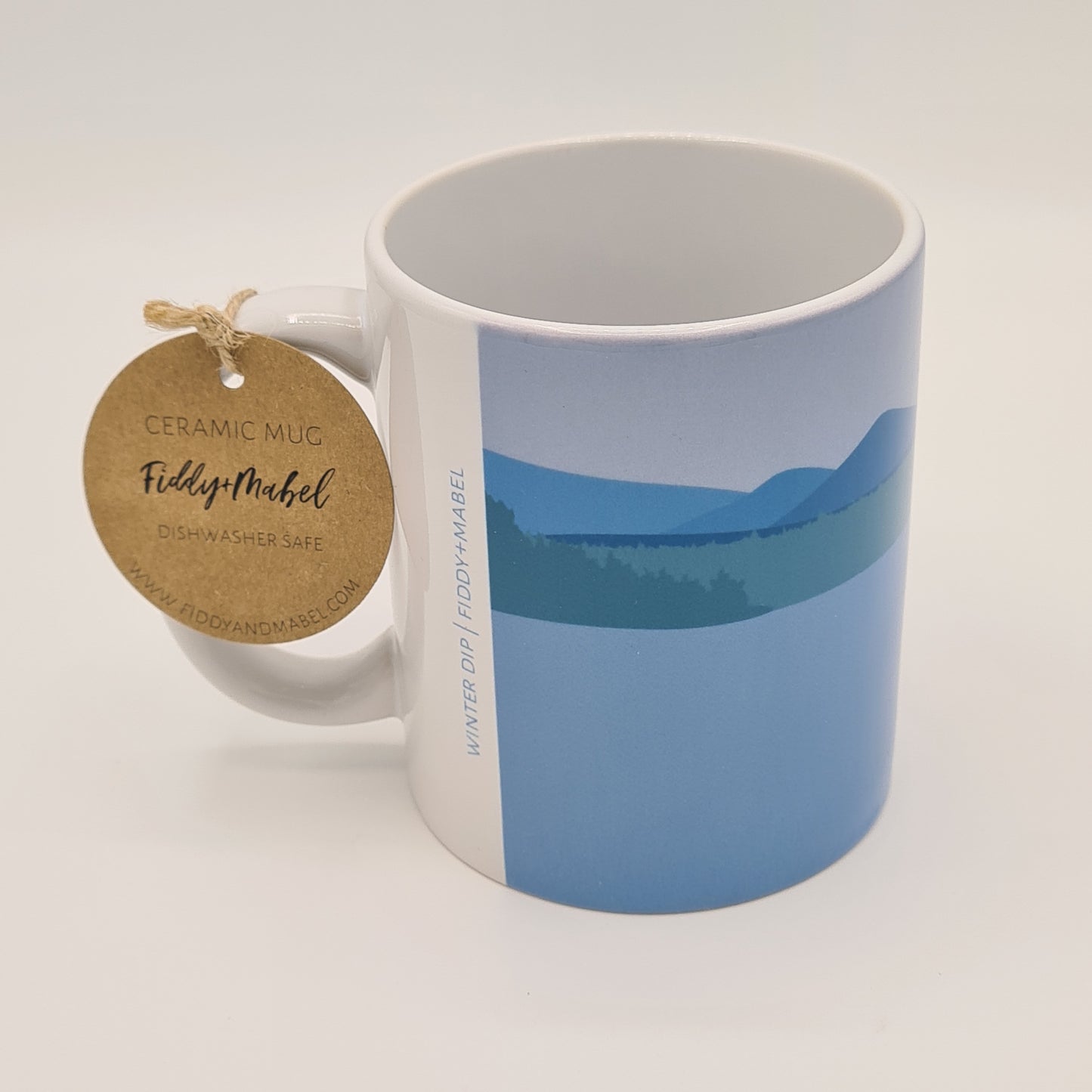 Side view of mug from Fiddy & Mabel entitled "Winter Dip". It shows an illustration of the water, a grass bank and hills/mountains. There is a round brown label tied to the handle of the mug stating "Ceramic Mug" "Dishwasher Safe" and it also features the logo for Fiddy & Mabel and their web address. 