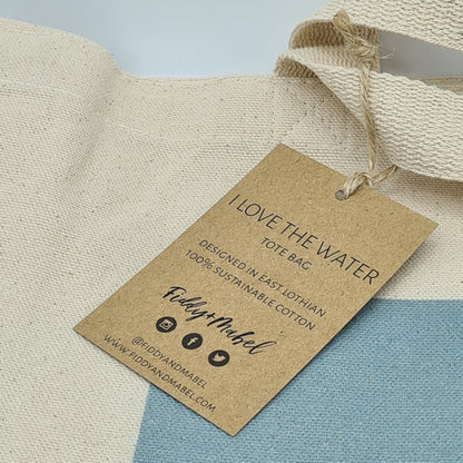 Close up of the label for the tote bag. It is a brown label with the text "I Love the Water tote bag"; "Designed in East Lothian, 100% sustainable cotton". It features the logo for Fiddy & Mabel, their website address and icons for Instagram, Facebook and Twitter.