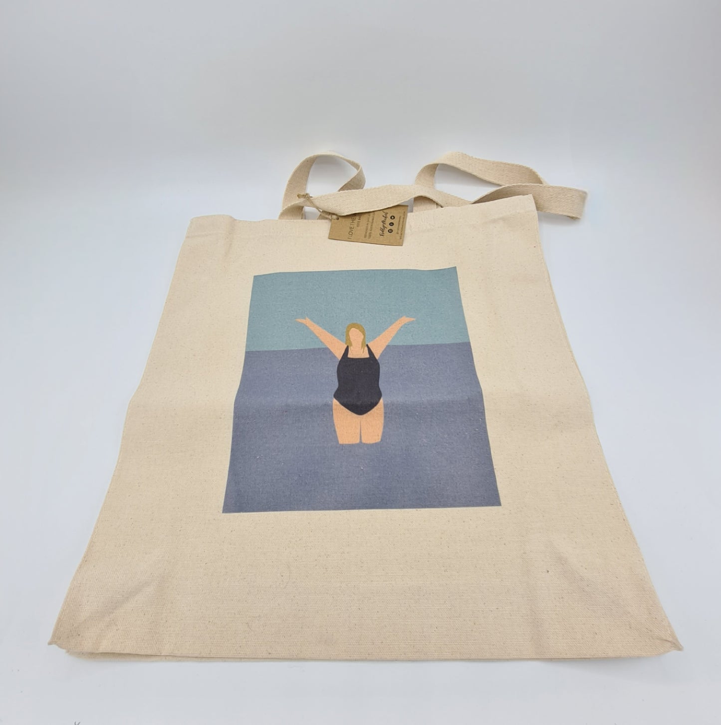 Cream coloured tote bag from Fiddy & Mabel entitled "I Love the Water". It features an illustration of a woman standing in the sea. She is wearing a dark swimsuit and holding her arms up high in joy.