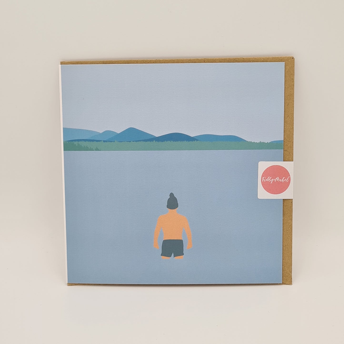 Front of greetings card from Fiddy & Mabel entitled "Winter Dip". It features an illustration of a man from the rear. He is wearing swimming shorts and a woollen hat and is standing in the water looking towards a grass bank and hills/mountains.