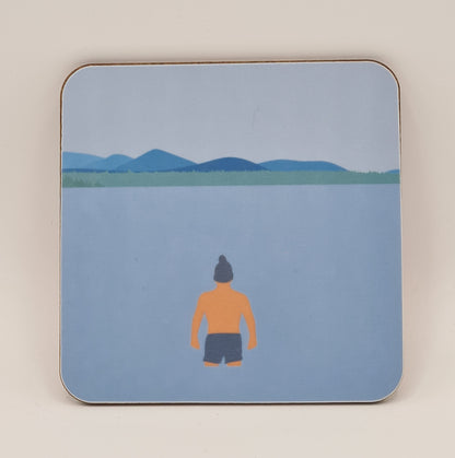 Square coaster from Fiddy & Mabel entitled "Winter Dip". It features an illustration of a man from the rear. He is wearing swimming shorts and a woollen hat and is standing in the water looking towards a grass bank and hills/mountains.