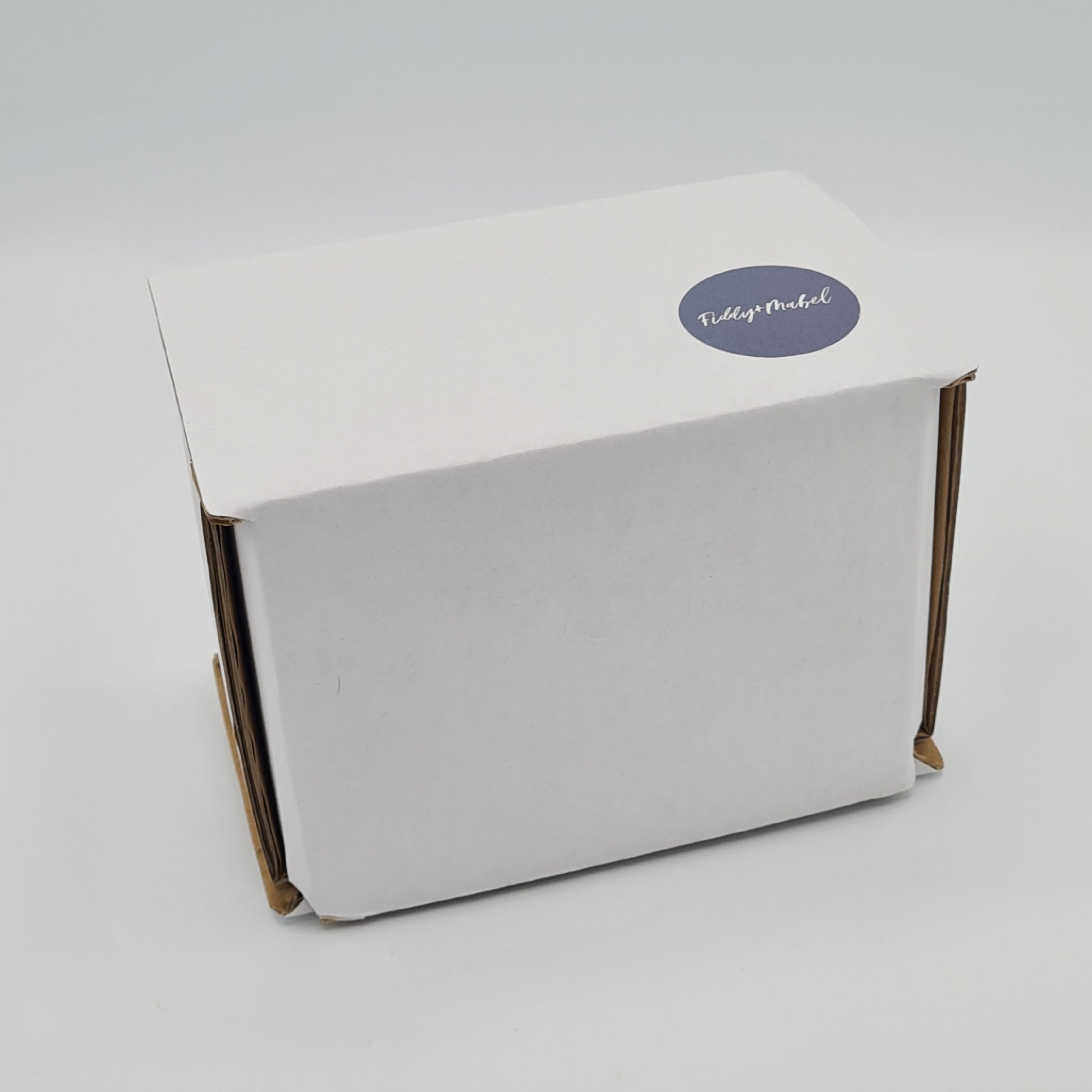 White box which is the packaging for the ceramic mug from Fiddy & Mabel. It features a label with their logo on it.