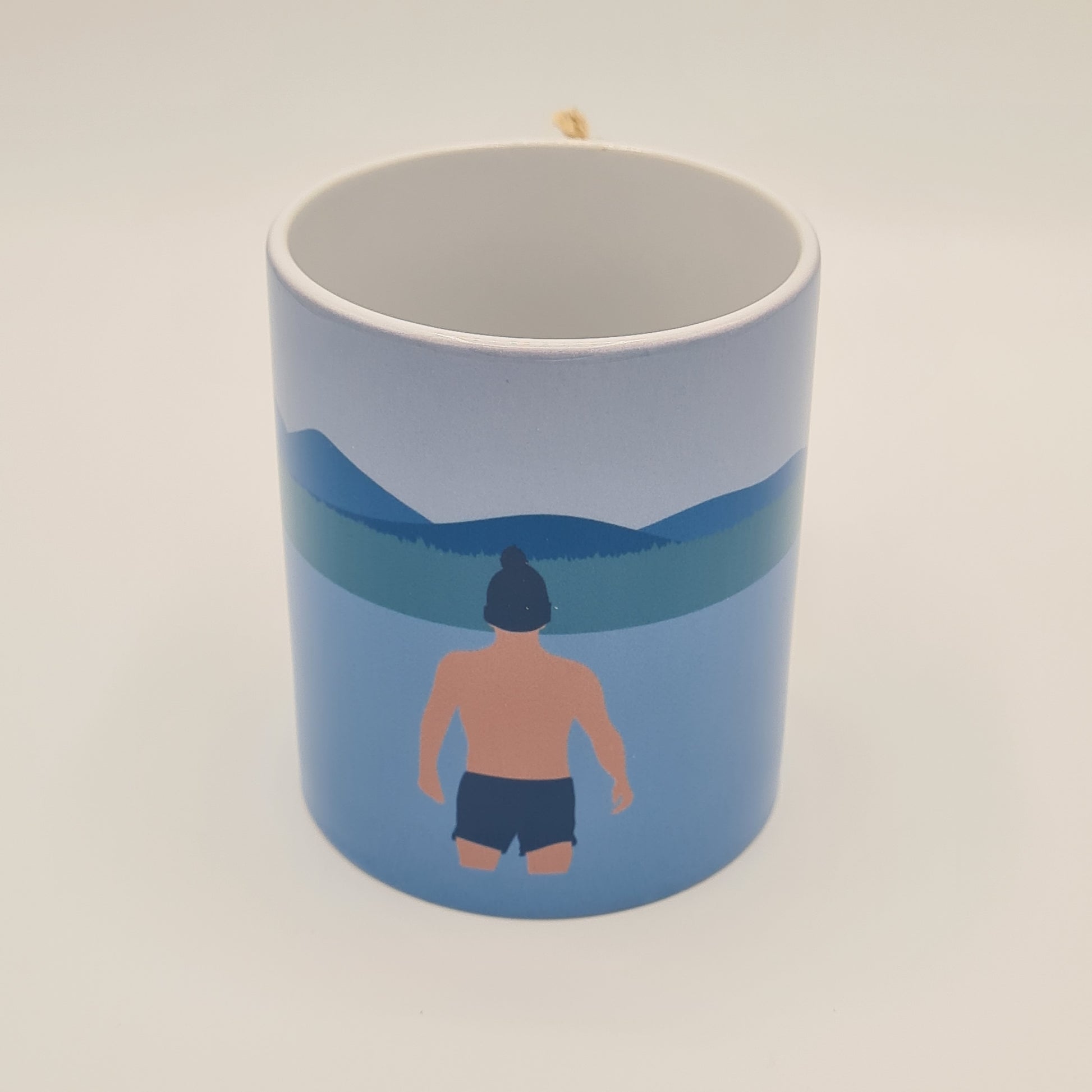 Mug from Fiddy & Mabel entitled "Winter Dip". It features an illustration of a man from the rear. He is wearing swimming shorts and a woollen hat and is standing in the water looking towards a grass bank and hills/mountains.