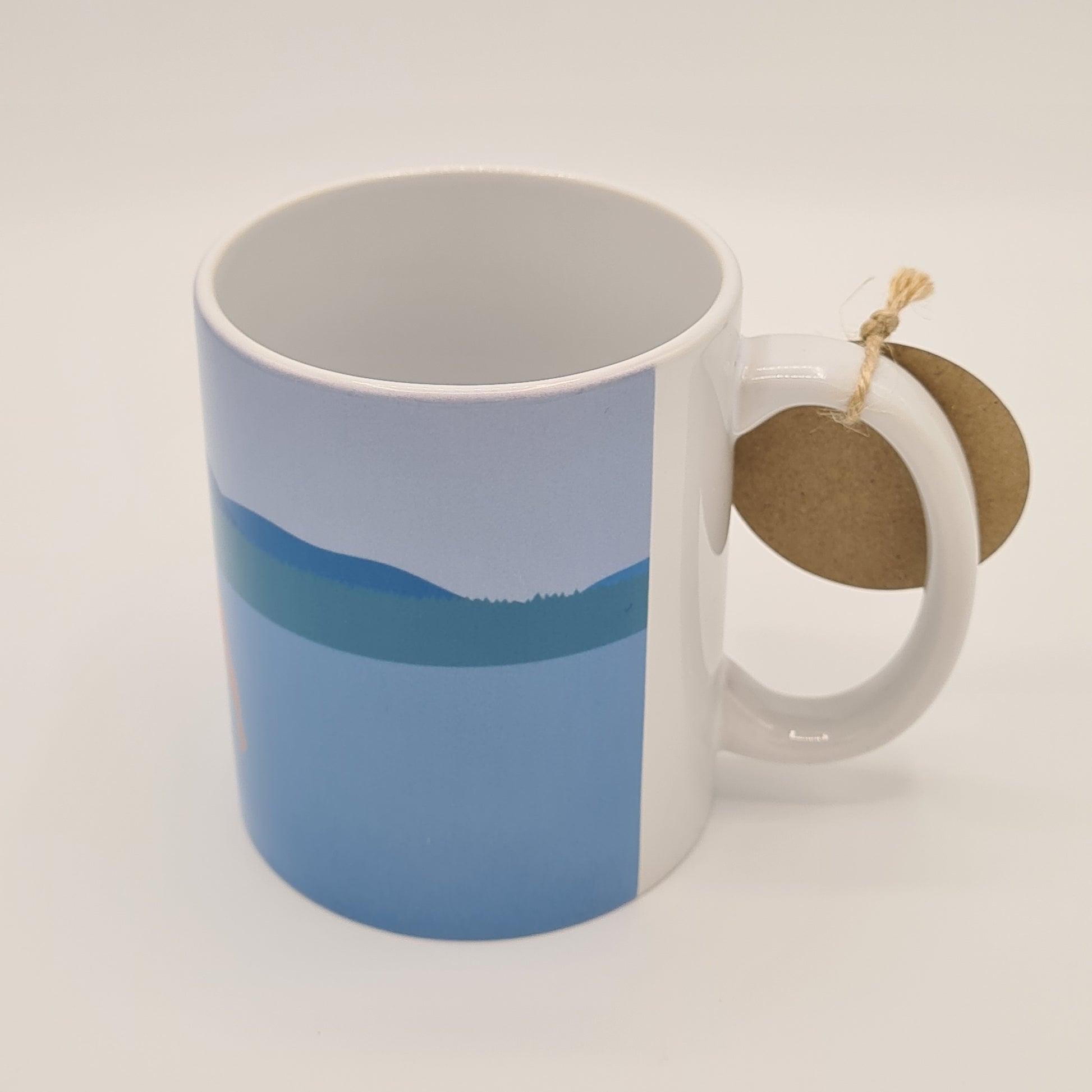 The other side of the mug from Fiddy & Mabel entitled "Winter Dip". It features an illustration of water, grass bank and hills/mountains. The handle of the mug is white.