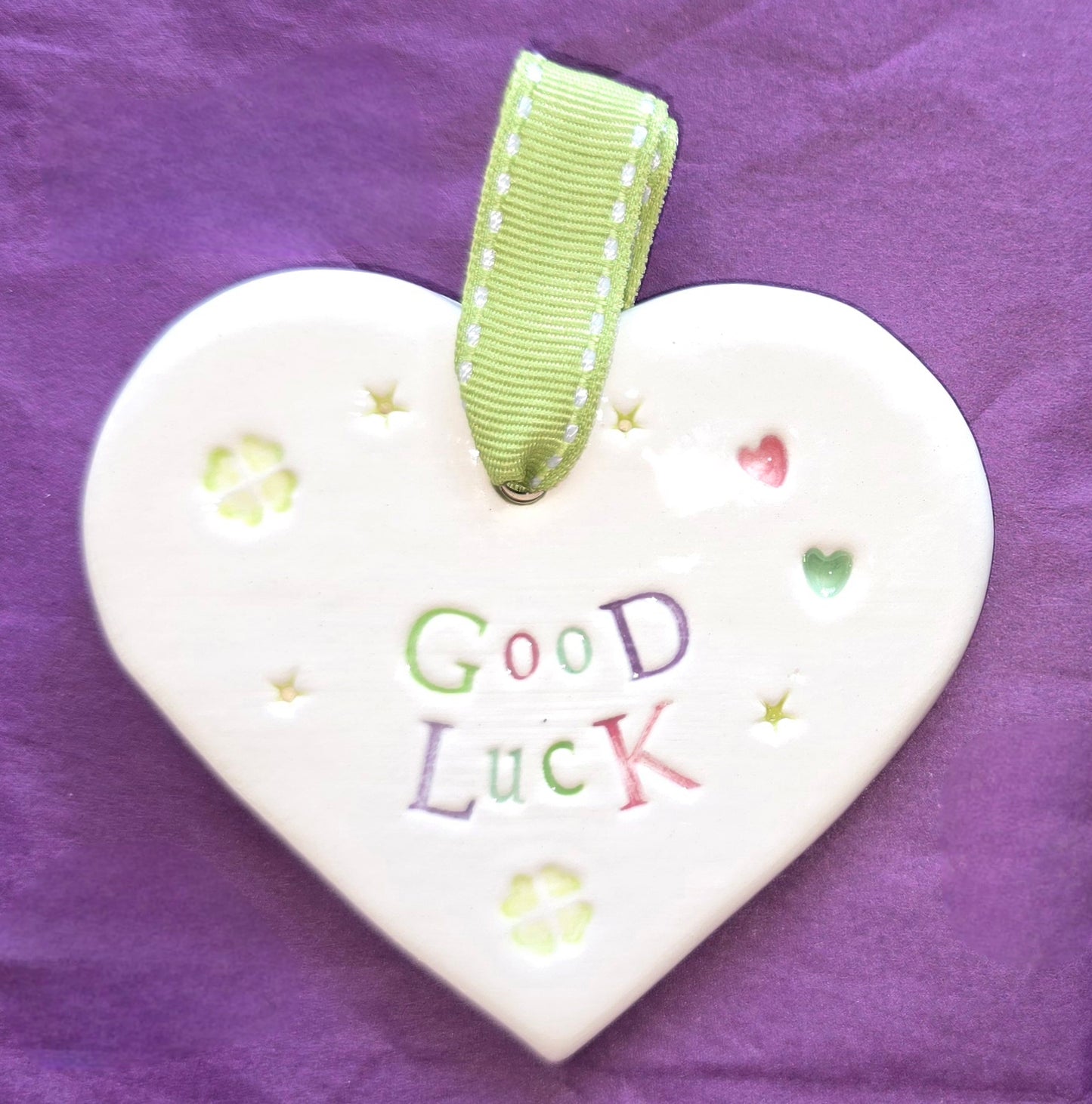 White ceramic heart with the words "Good Luck" and some small hearts and clover illustrations dotted around. There is a green ribbon at the top of the heart for hanging.