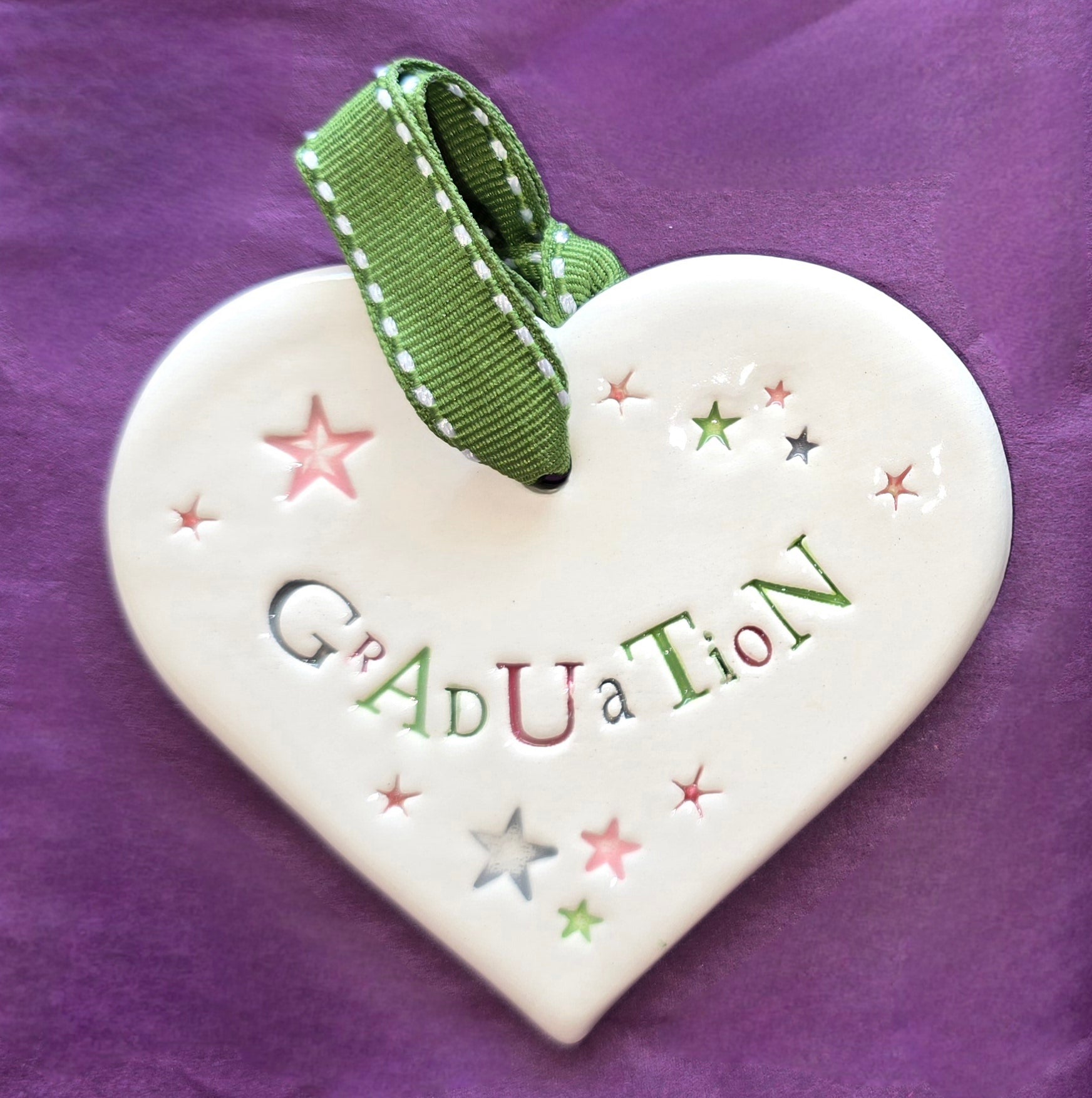 White ceramic heart with the words "Graduation" and some small star shaped illustrations dotted around. There is a green ribbon at the top of the heart for hanging.