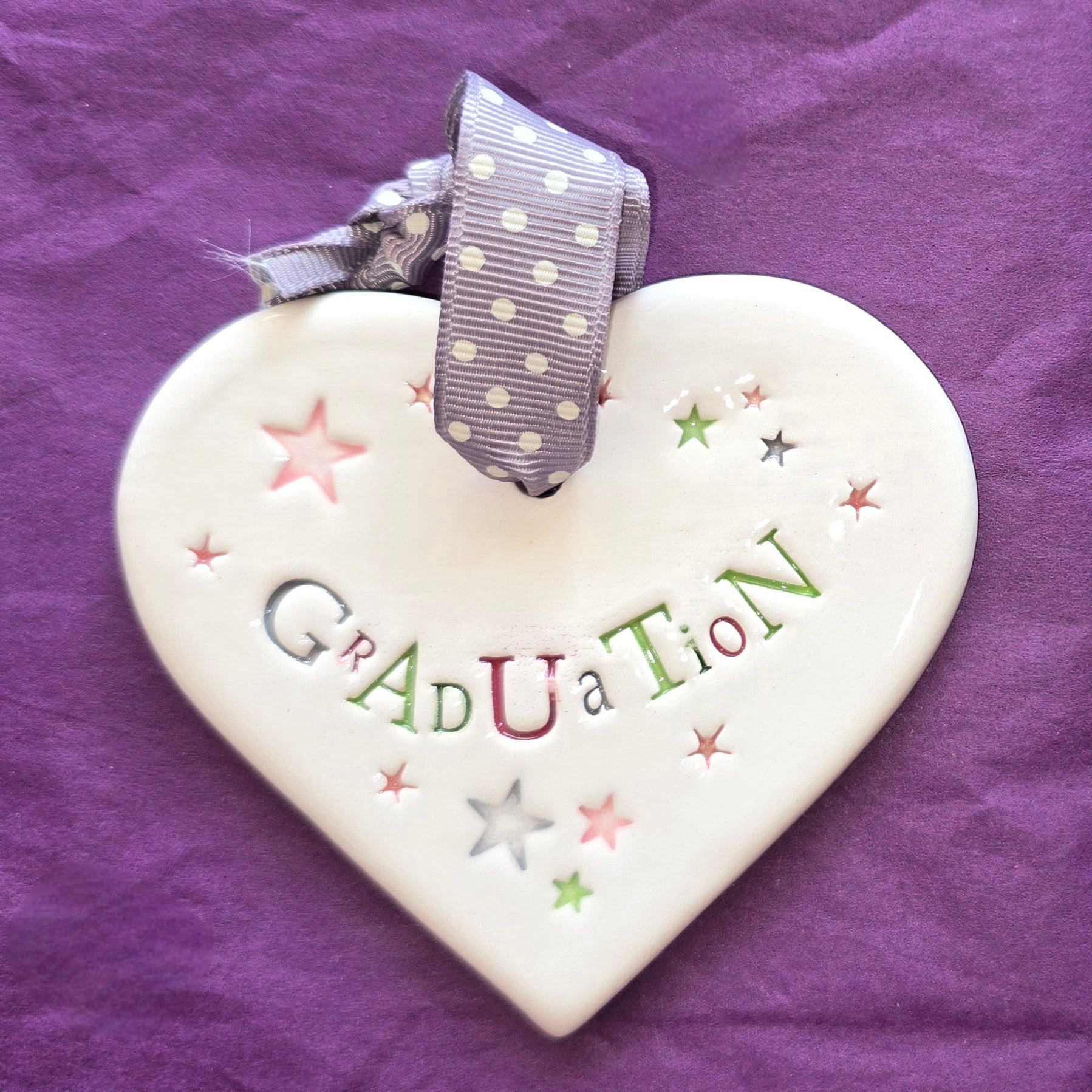 White ceramic heart with the words "Graduation" and some small star shaped illustrations dotted around. There is a grey polka dot ribbon at the top of the heart for hanging.