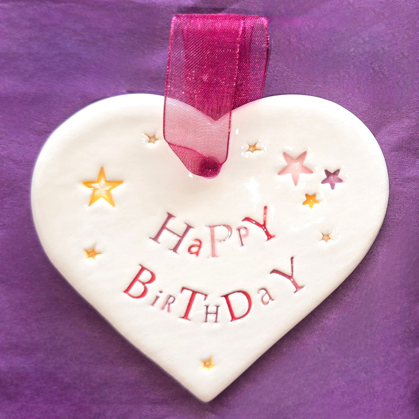 White ceramic heart with the words "Happy Birthday" and some small star shaped illustrations dotted around. There is a burgundy ribbon at the top of the heart for hanging.